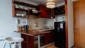 A compact kitchen with wooden cabinets, a black refrigerator, a microwave, and a hanging light fixture. - Rbk Moderno dpto ubicación inmejorable (Santiago)