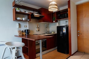 Eine kompakte Küche mit Holzschränken, einem schwarzen Kühlschrank, einer Mikrowelle und einer Pendelleuchte.