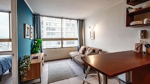 A modern living room with a sofa, a dining table, a plant, and a large window with a city view. - Rbk Moderno dpto ubicación inmejorable (Santiago)