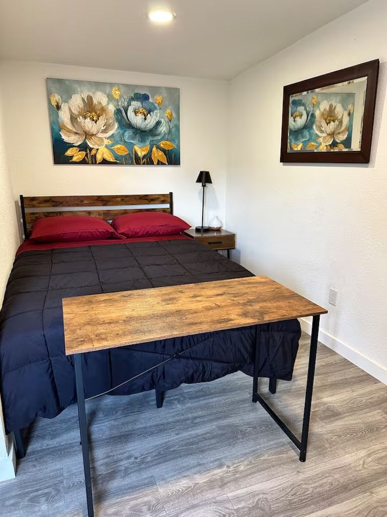 A bedroom with a bed, a wooden bedside table, a nightstand, and two paintings on the wall.