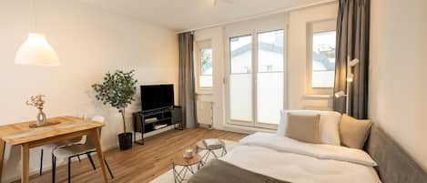 A modern hotel room with a wooden desk, a flat-screen TV, a bed, and large windows.