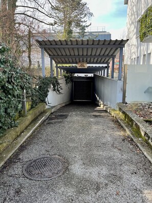 Une entrée abritée, avec un chemin de gravier menant à une zone délimitée par une grille, entourée de verdure et avec un bâtiment en arrière-plan.