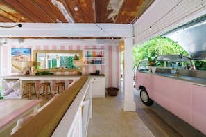 Ein pinkfarbener Van, umgebaut zu einem Food-Truck mit Verkaufstresen und Hockstühlen, rustikaler Decke und Stadtbild-Gemälde.