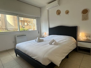 Ein ordentlich bezogenes Bett mit weißen Bettwäsche und Handtüchern, ein Nachttisch mit Lampe sowie ein Fenster mit Blick auf Gebäude.