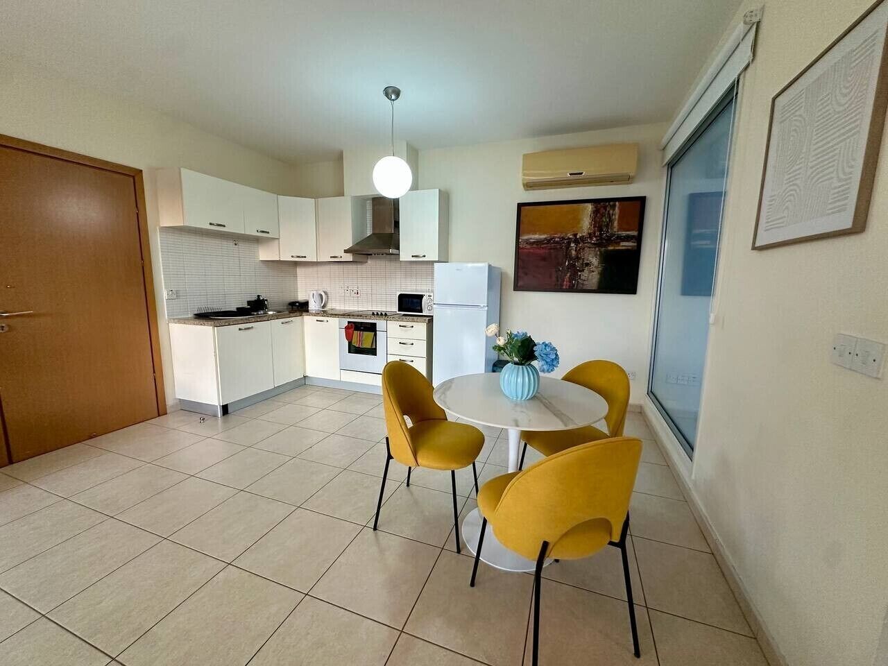 A compact kitchen with white cabinets, a dining area with yellow chairs, and a large window.