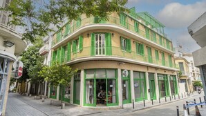 A multi-story building with green shutters, a curved facade, and a ground-level shop with a sign indicating it is for rent. - Renovated city center studio balcony/terrace view (Nicosia)