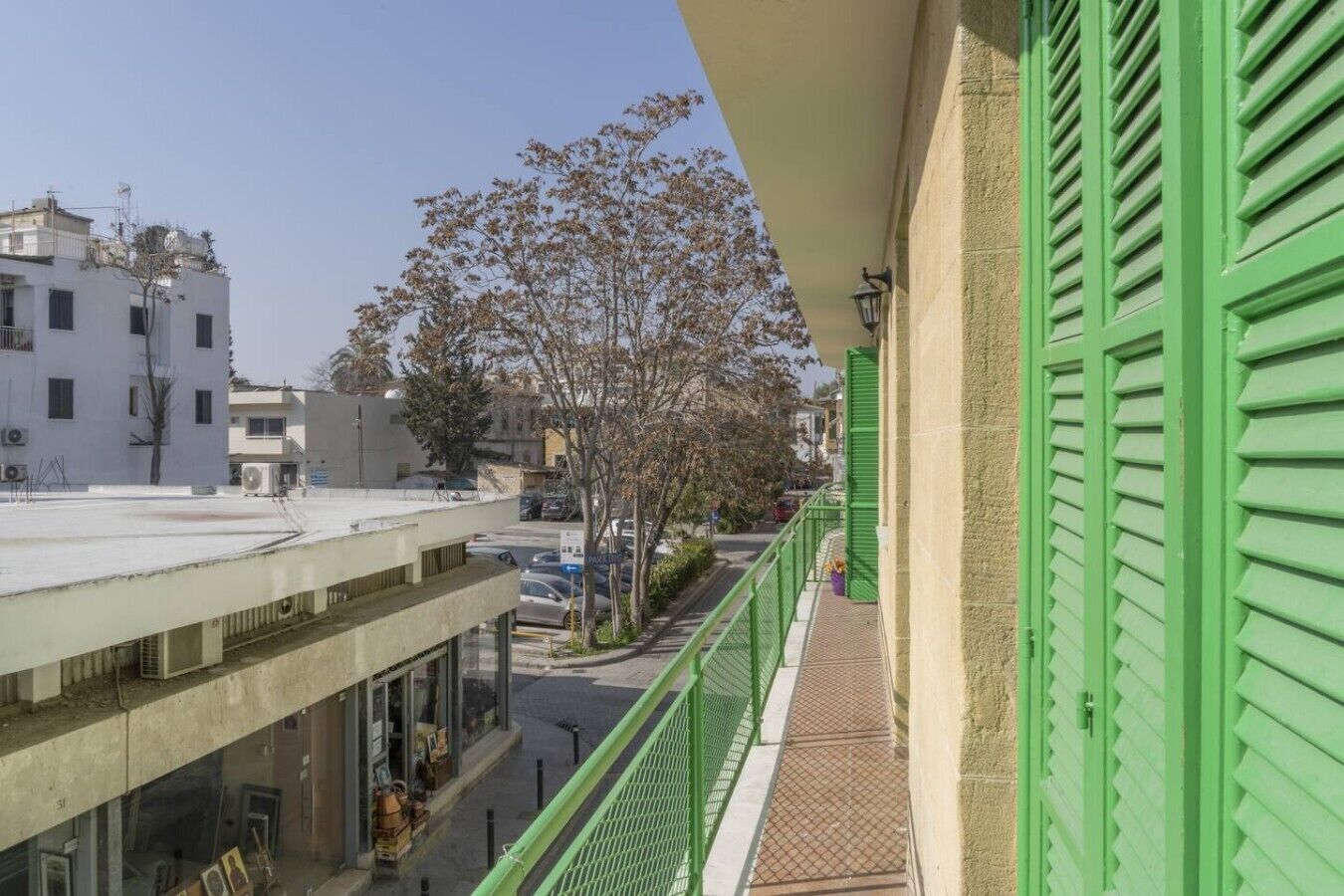 Un balcón con contraventanas verdes que da a una calle con coches estacionados y edificios.