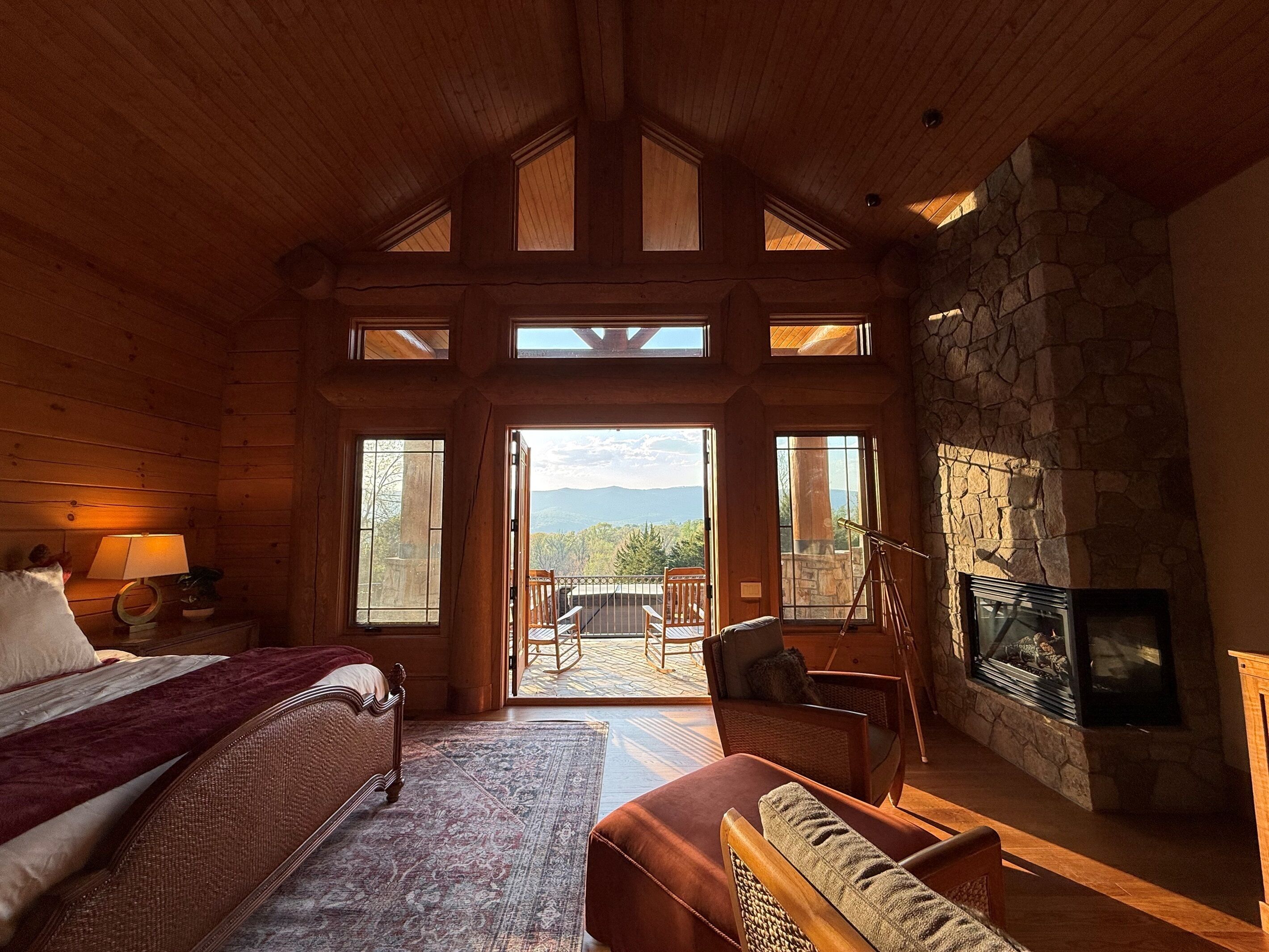 A log cabin interior with a bed, a fireplace, a stone wall, and a balcony with a view of mountains.
