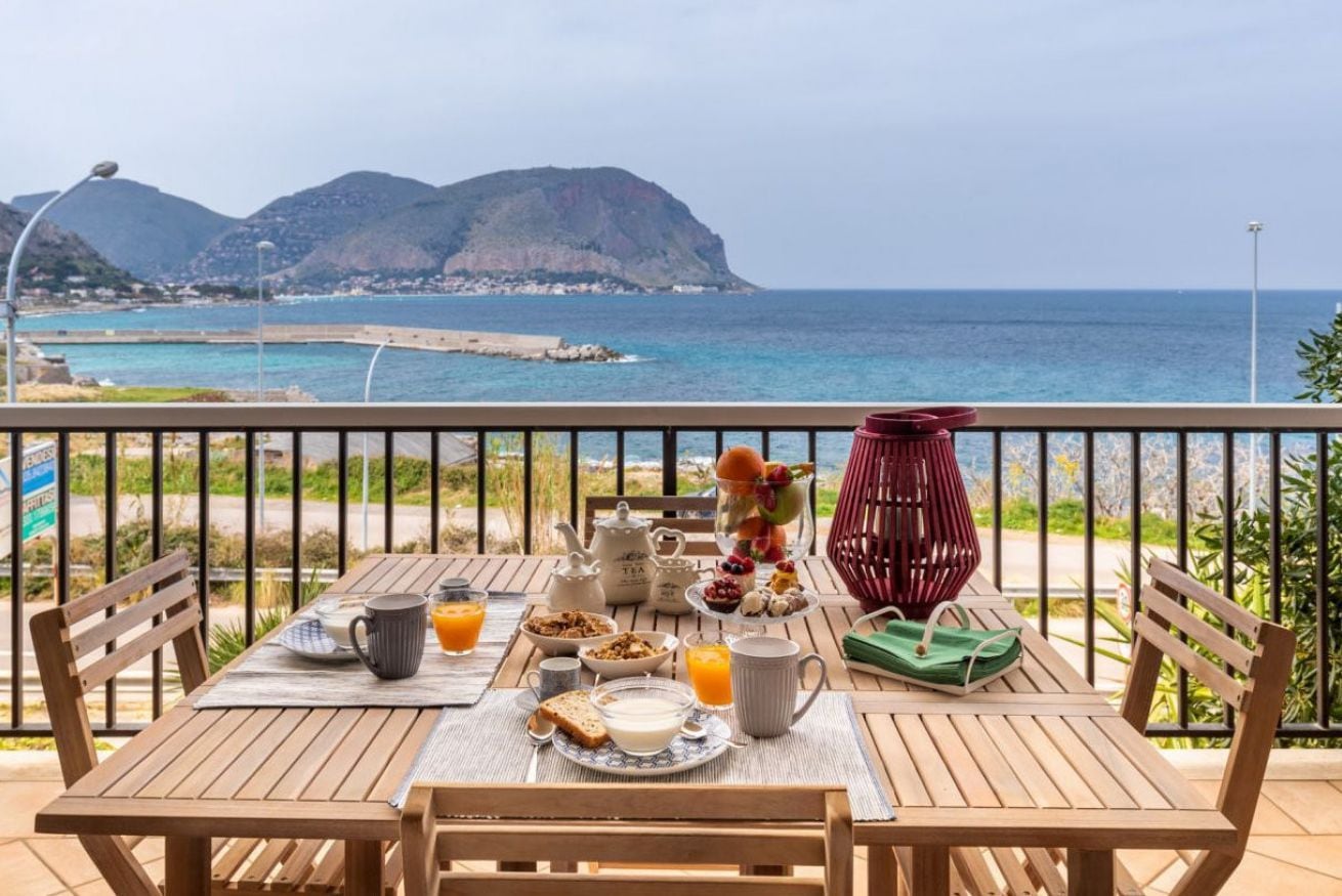 A wooden table with breakfast items, a view of the sea, and a mountainous landscape.