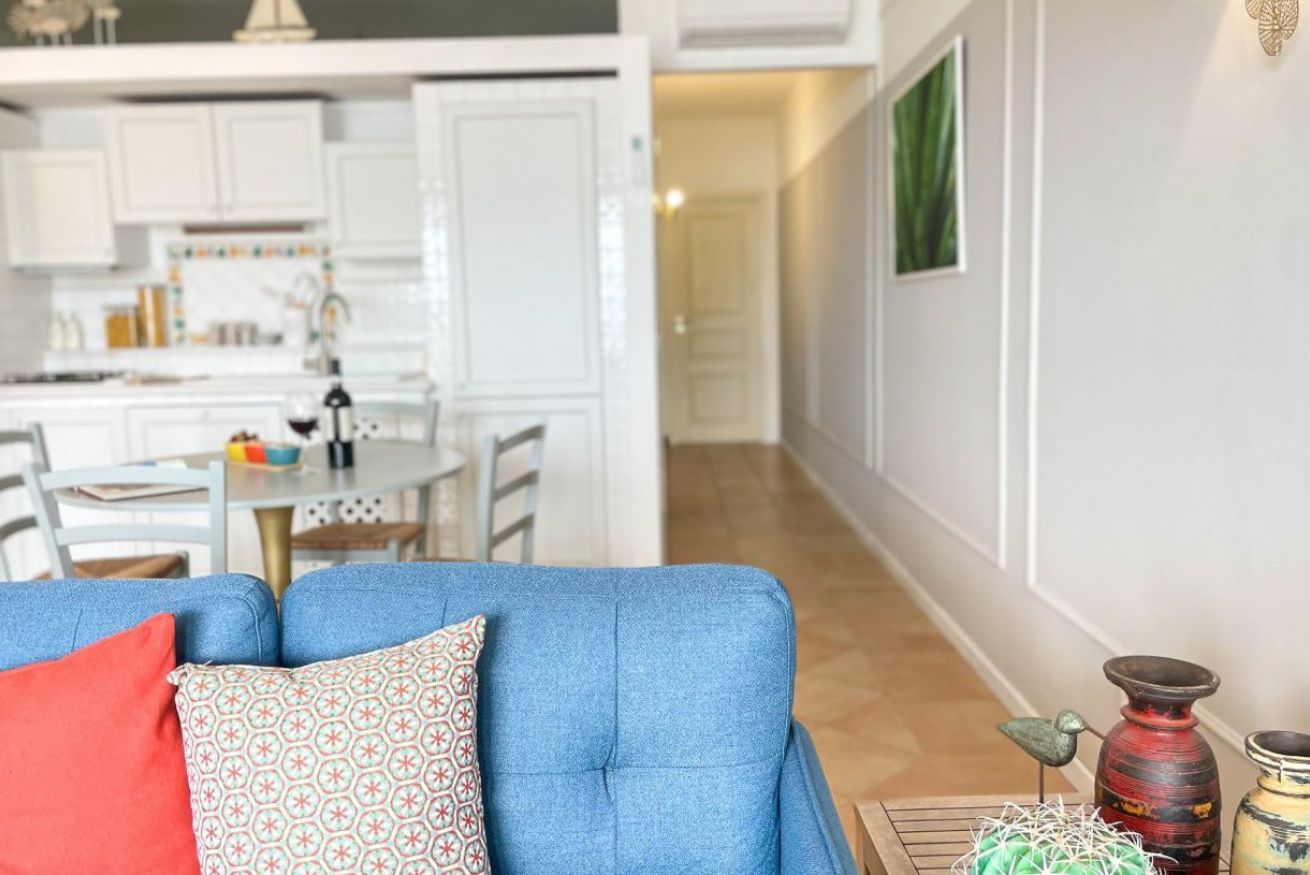 A living room with a blue sofa, a dining area with white cabinets, and a hallway leading to another room.