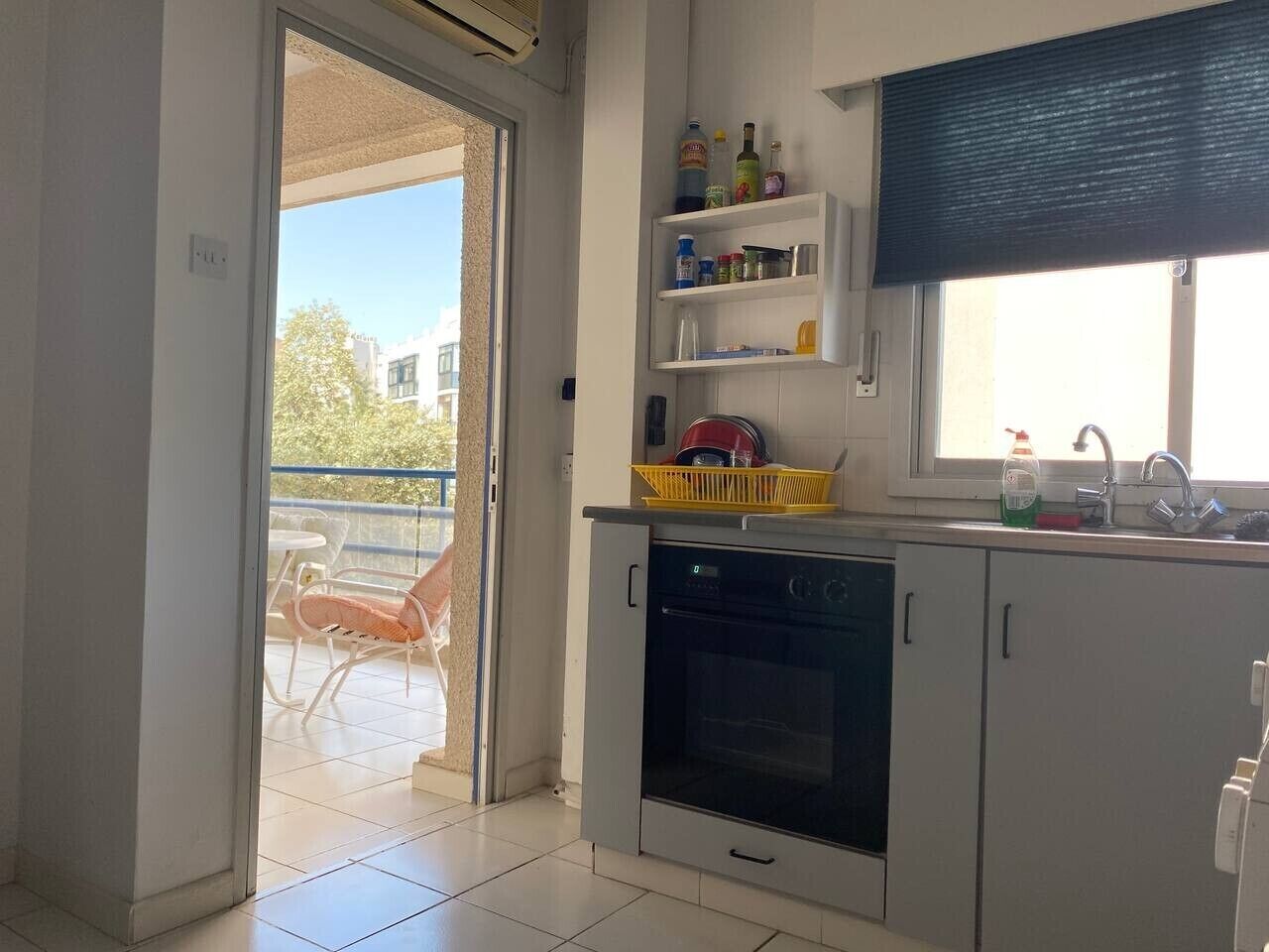 A kitchen with a built-in oven, a sink, and a window with a blind. There is a balcony with a chair visible through the door.