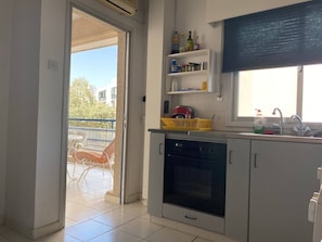 A kitchen with a built-in oven, a sink, and a window with a blind. There is a balcony with a chair visible through the door. - Private single room in shared flat, best location (Limassol)