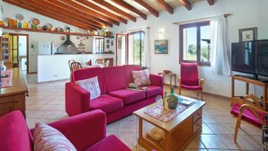 Una sala de estar acogedora con un sofá rojo, una mesa de centro de madera y un televisor. Se ve una cocina con vigas de madera y una zona de comedor con sillas y una mesa al fondo.