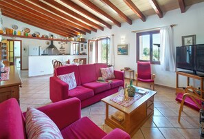 A cozy living room with a red sofa, wooden coffee table, and a television. A kitchen with wooden beams and a dining area with chairs and a table are visible in the background. - Tranquil Countryside Finca, A/C Private Pool - 15 mins drive from Pollenca, Alcudia & Beaches (Alcudia)