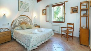 Un dormitorio con cama, una silla, mesita de noche, estante y ventana.