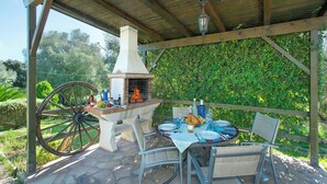 Un patio exterior con un horno de ladrillo, una mesa de madera lista para comer y una rueda de carro antigua.