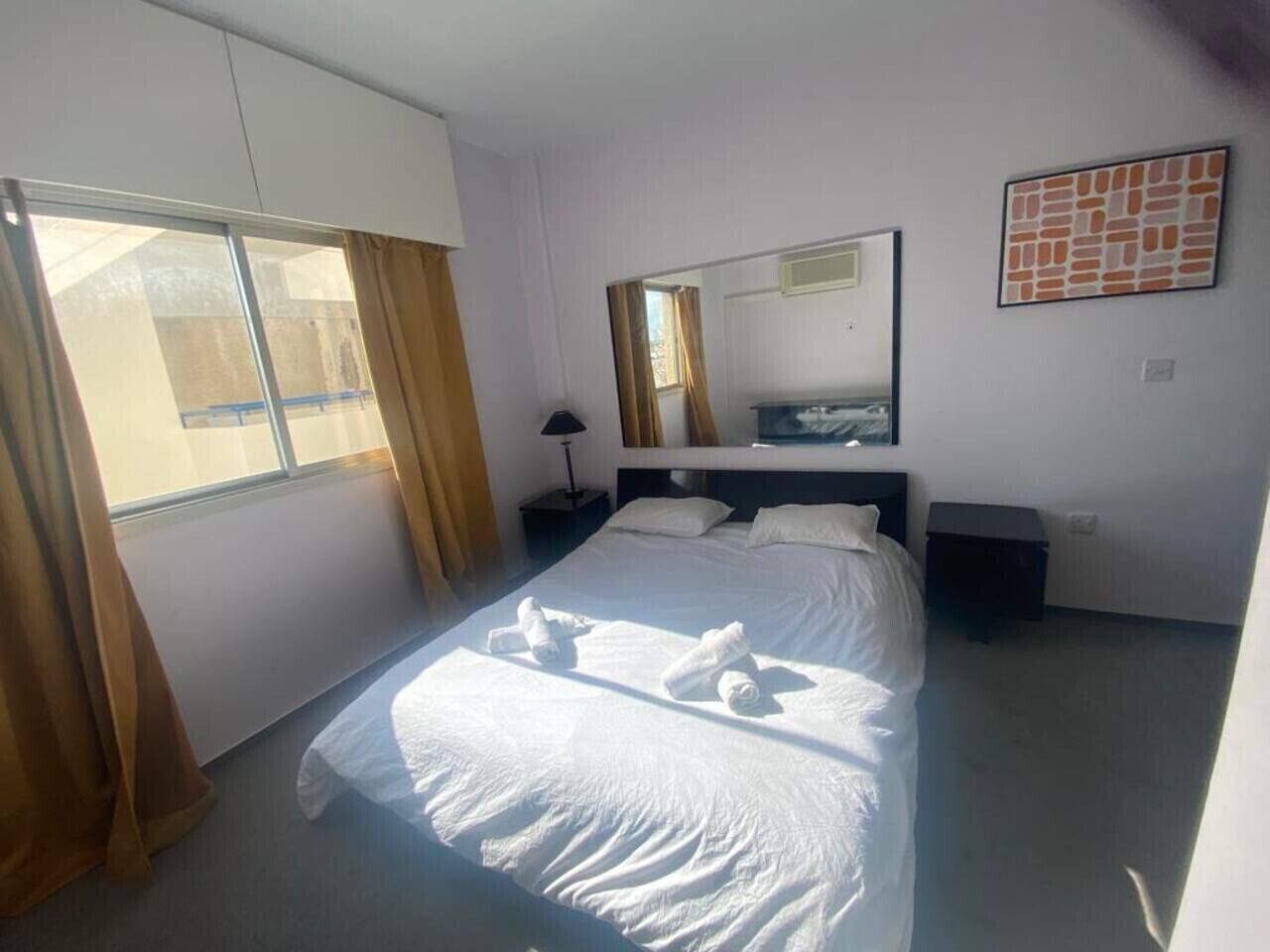 A hotel room with a bed, a nightstand, a lamp, a window with curtains, and a mirror on the wall.