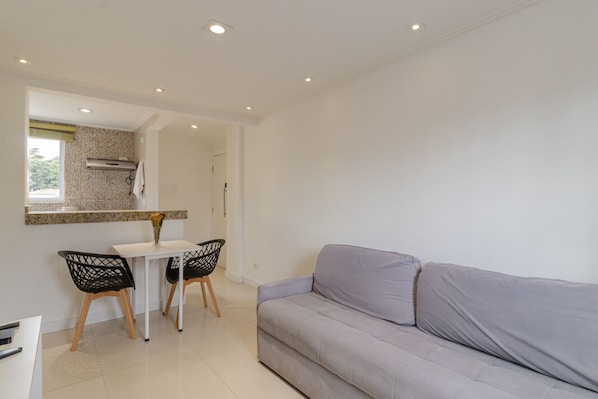 Ein modernes Wohnzimmer mit einem grauen Sofa, einem Essbereich mit einem weißen Tisch und zwei Stühlen sowie einer im Hintergrund sichtbaren Küche.
