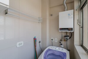 Ein Badezimmer mit weiß gefliesten Wänden, einem weißen Boiler, einer Waschmaschine und einem Fenster.