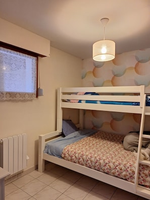 Une chambre avec un lit superposé, un radiateur, une fenêtre ornée de rideaux en dentelle et un mur décoré d’un motif circulaire.