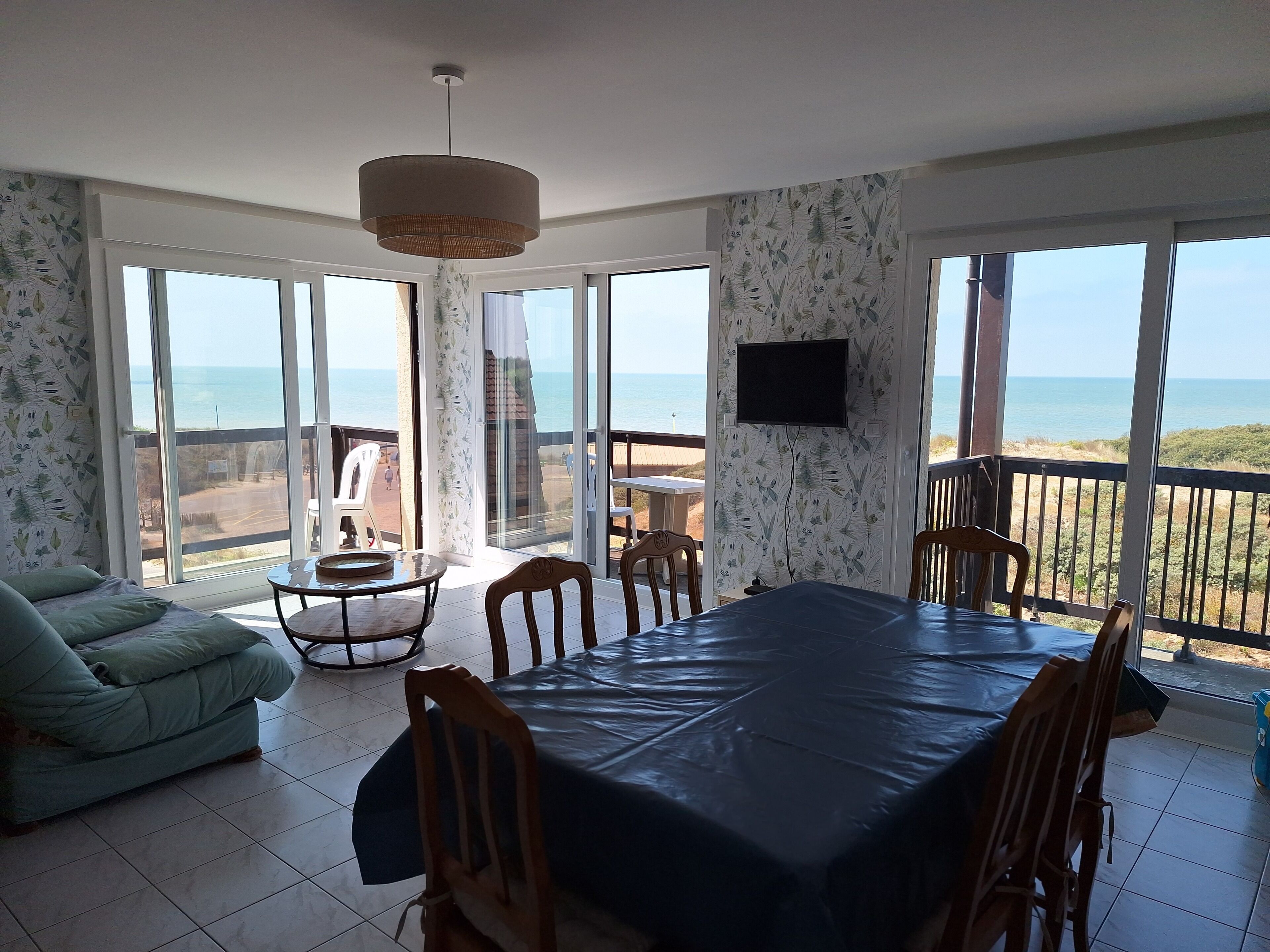 Une pièce comprenant une table à manger, des chaises, un canapé, un téléviseur et de grandes fenêtres donnant sur la mer.