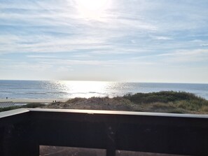 Une vue sur la plage depuis un balcon doté d’une balustrade, avec l’océan et un ciel dégagé en toile de fond.
