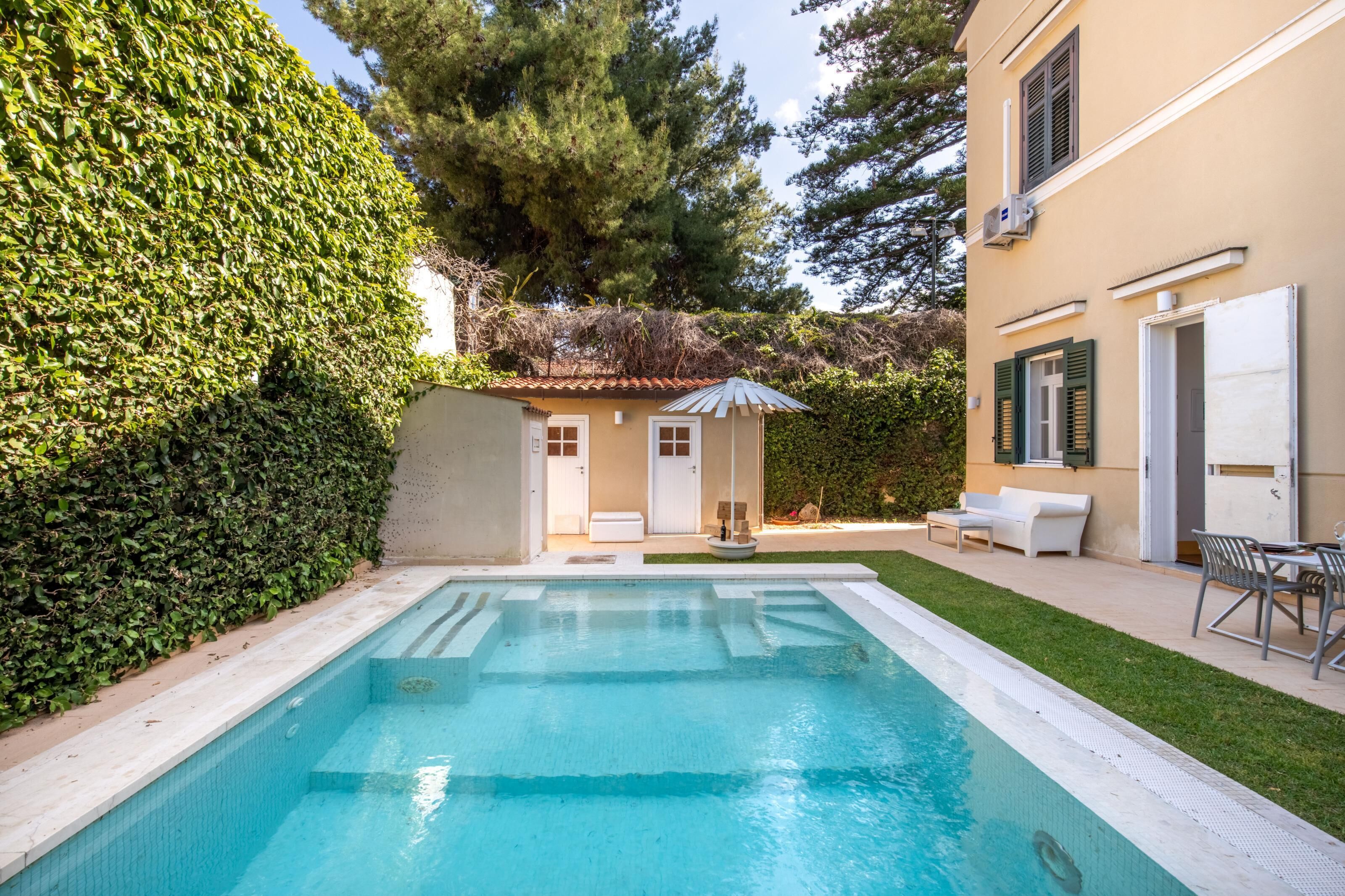 Une piscine avec une terrasse carrelée, un petit espace patio avec un canapé et une table blancs, et une haie qui entoure la propriété.