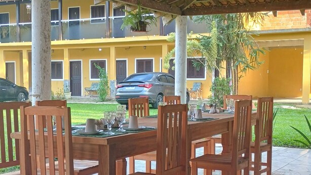 A covered outdoor dining area with wooden tables and chairs, a tiled floor, and a yellow building in the background. - Pousada cachoeira  (Paraty)
