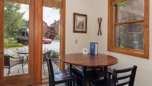 Een kamer met een ronde tafel, twee stoelen, een lijst met afbeelding en een raam met uitzicht op een straat en gebouwen.