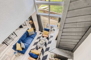 Ein Wohnbereich mit hohen Decken, einem blauen Sofa, gemusterten Kissen, einem Couchtisch und Blick auf eine Holzterrasse und einen Garten.