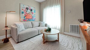 Ein Wohnzimmer mit einer Couch, einem runden Couchtisch, einer Lampe, einem Gemälde und einem Fenster mit Vorhängen.