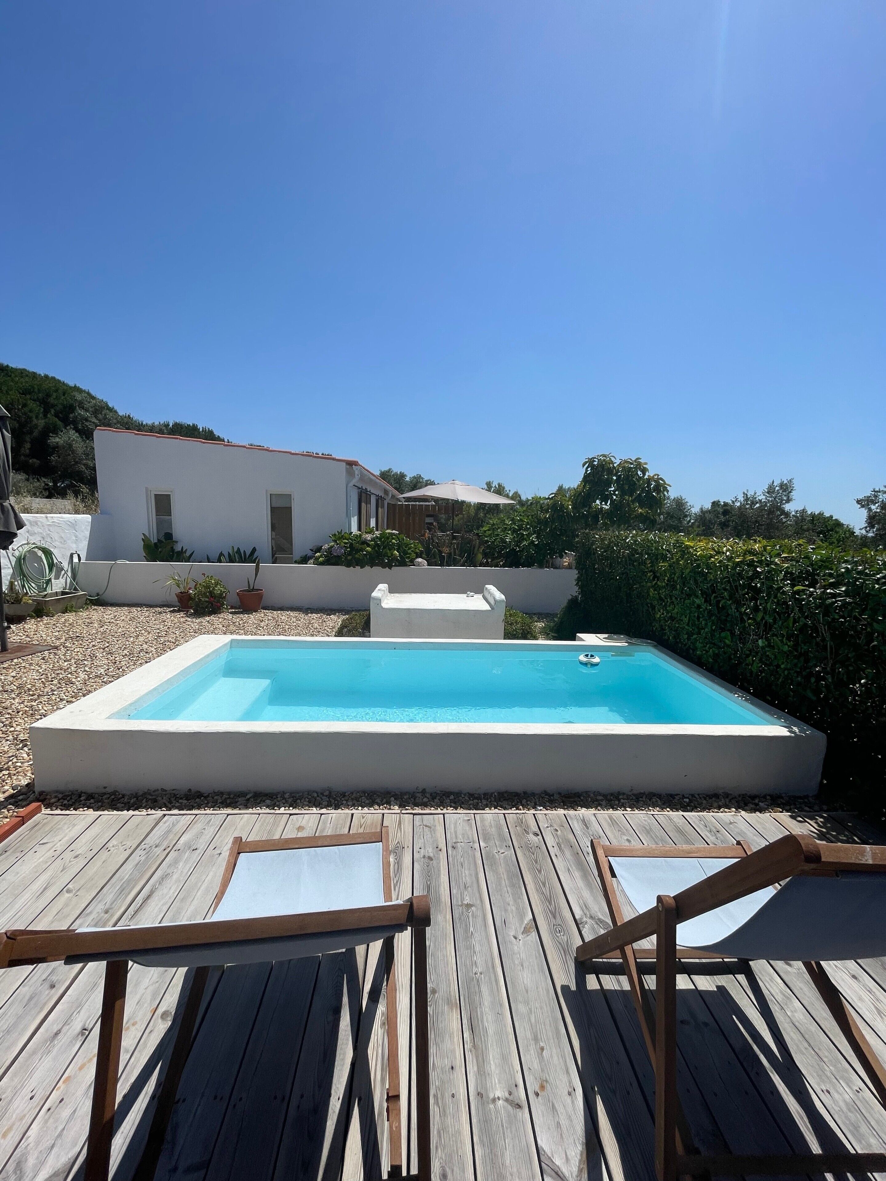Una piscina rectangular con muelle de madera al frente, un edificio blanco al fondo y un cielo azul despejado.