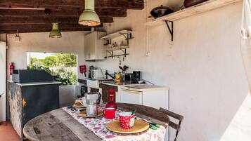 A rustic kitchen with wooden beams, a dining table with a floral tablecloth, and a view of greenery outside.