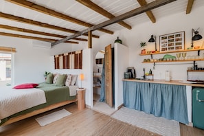 A compact bedroom with a bed, bedside table, and a nightstand. There is a kitchenette area with a sink, stove, and shelves above.