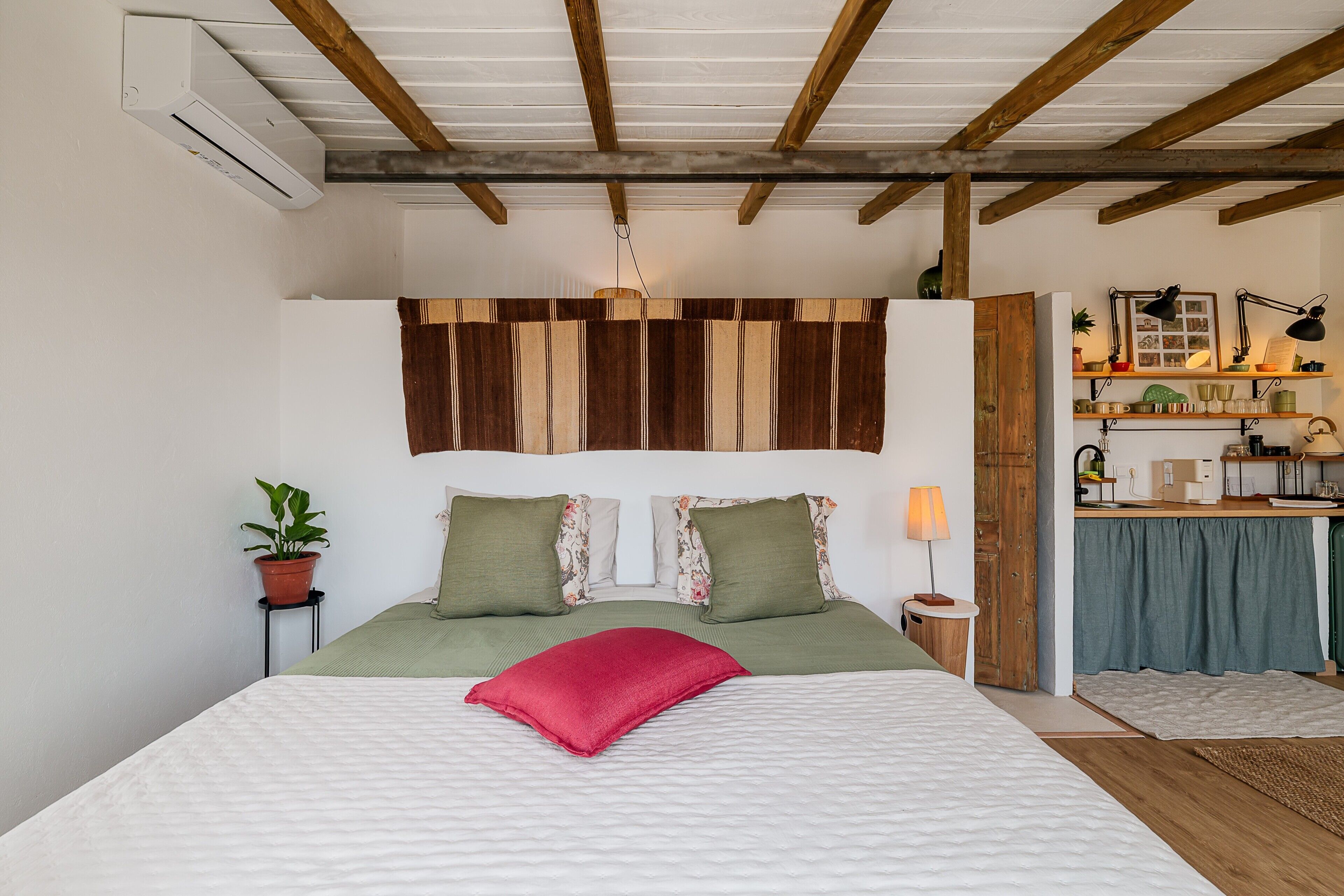 A bedroom with a bed, a nightstand, a lamp, a potted plant, and a wooden ceiling.