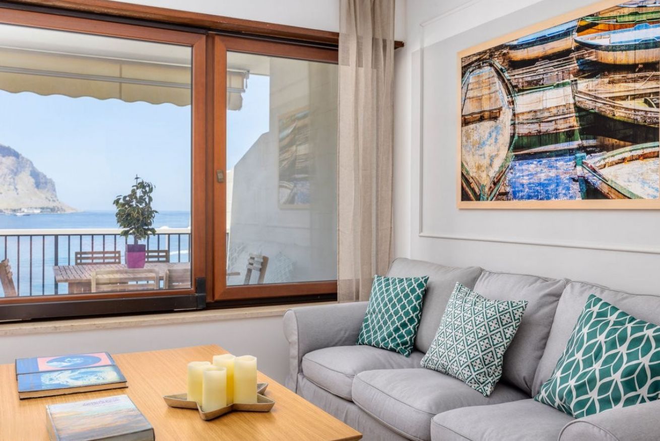 Een kamer met een grijze bank, decoratieve kussens, een houten tafel met kaarsen, een kunstwerk in een lijst en een balkon met uitzicht op zee en een klif.