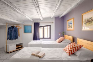 Ein Zimmer mit zwei Betten, einem Holzkopfstück, einem Fenster mit Vorhängen, einem Kleiderständer mit Kleidung und einer Wand mit gerahmten Bildern.