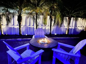 An outdoor seating area with a round table and chairs, a fire pit, and a white picket fence with palm trees. - 
The perfect getaway ,ONLY 2 Minutes away from  the beach!!!! (Fort Lauderdale)