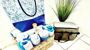 Un cesto di vimini con asciugamani, una boccetta di crema solare, una borsa blu e una valigia grigia su un pavimento piastrellato.