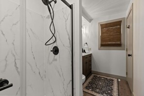 Un baño con ducha, inodoro y lavabo, una alfombra estampada y una ventana con persianas.
