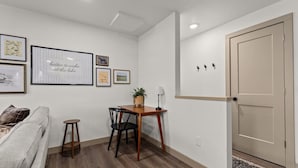 Habitació amb terra de fusta, un sofà, una taula de dinar amb cadires, una petita taula auxiliar amb una làmpada i una porta.