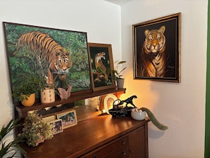 A wall with two framed tiger paintings, a wooden shelf with potted plants, a small cactus, and a decorative horse figurine.