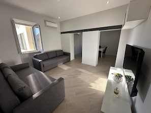 A modern living room with a grey sofa, a dining area with a white table, and a kitchen area with a white countertop.
