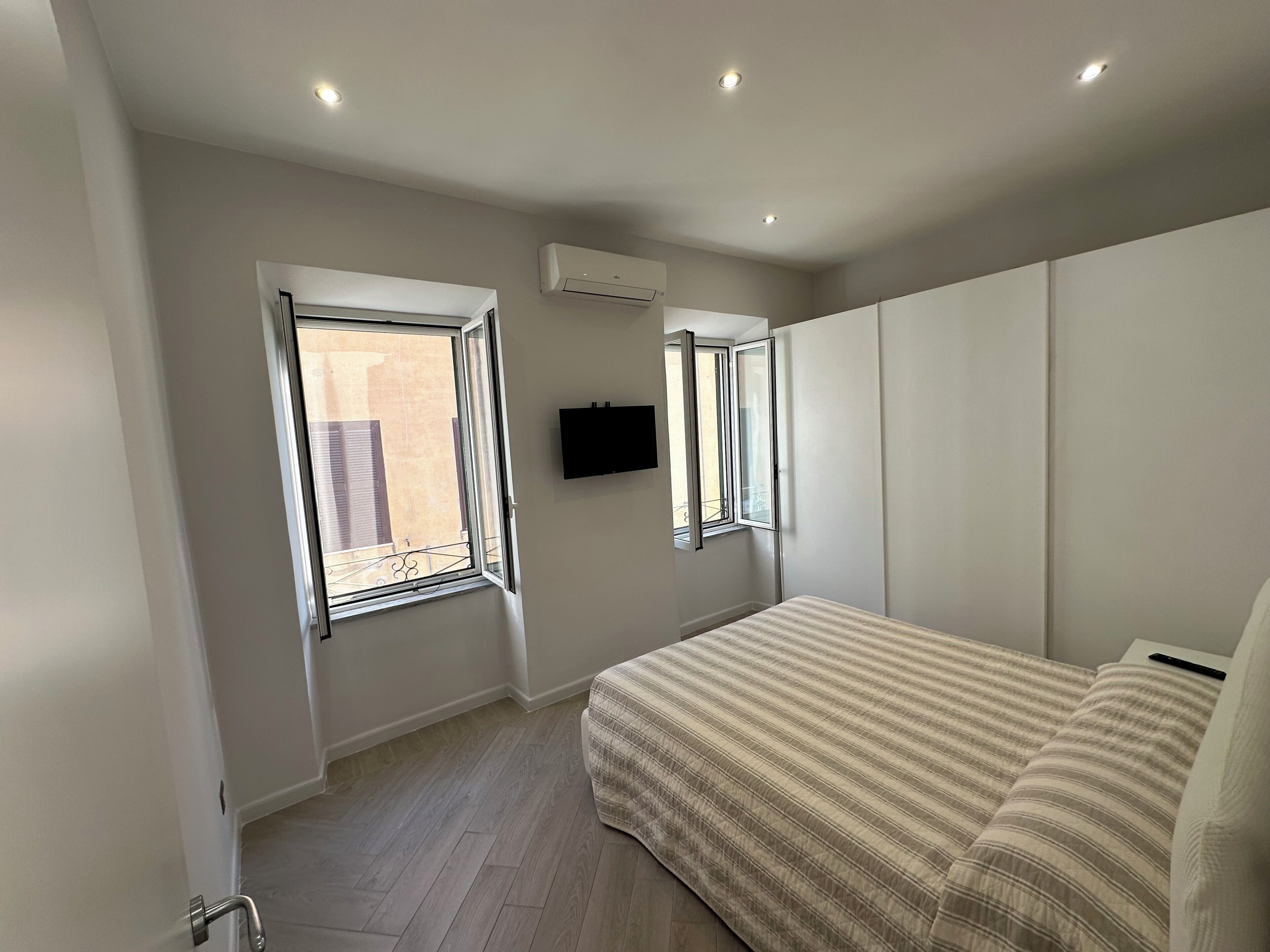Une chambre à coucher avec un lit, deux fenêtres, un climatiseur et un téléviseur fixé au mur.