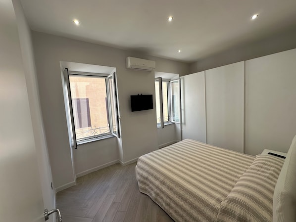 A bedroom with a bed, two windows, an air conditioning unit, and a wall-mounted TV.