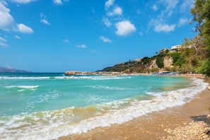 A beach with clear turquoise water, pebbly shore, and buildings on a hillside. - Bicorna Chania Pool Escape - Silver Room 3 (Apokoronas)