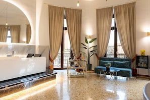 A modern hotel lobby with a reception desk, a sofa, a coffee table, and large windows with curtains. - Sanfelice Rooms & Suites (Naples)