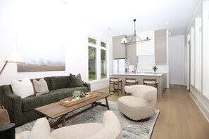 Ein modernes Wohnzimmer mit Sofa, Couchtisch und Sesseln, eine Küche mit Essbereich und großen Fenstern.