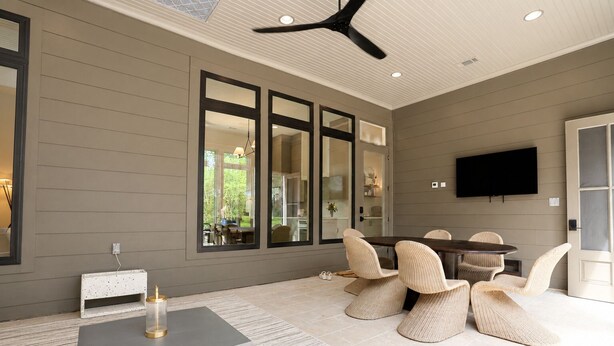 A modern living room with a grey wall, large windows, a ceiling fan, a TV, and a dining area with wicker chairs.