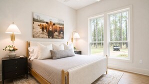 A bedroom with a bed, two bedside tables, a nightstand, a window with a view of trees and a vehicle, and a painting of cows.